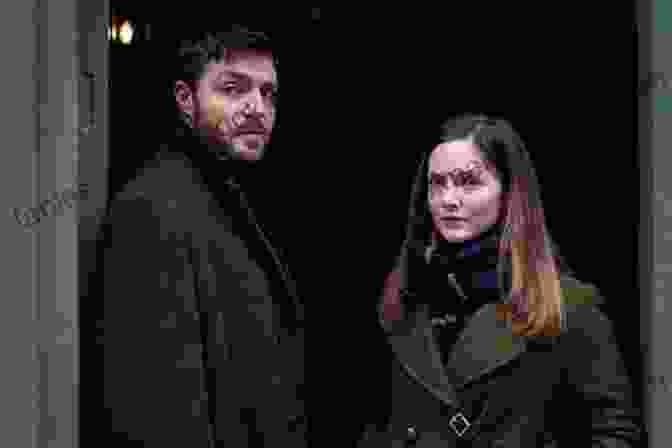 Cormoran Strike And Robin Ellacott, The Central Characters Of The Ink Black Heart, Standing In A Dimly Lit Room With Serious Expressions On Their Faces. The Ink Black Heart (A Cormoran Strike Novel 6)